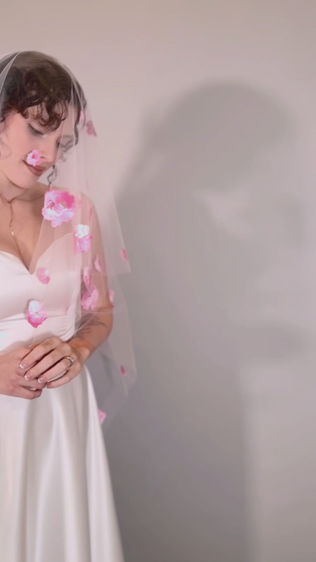 Woman in a white wedding dress with a long pink painted veil with pink floral petals against a plain background. Video by Taylor Mitchell Model is Isabelle Paulino