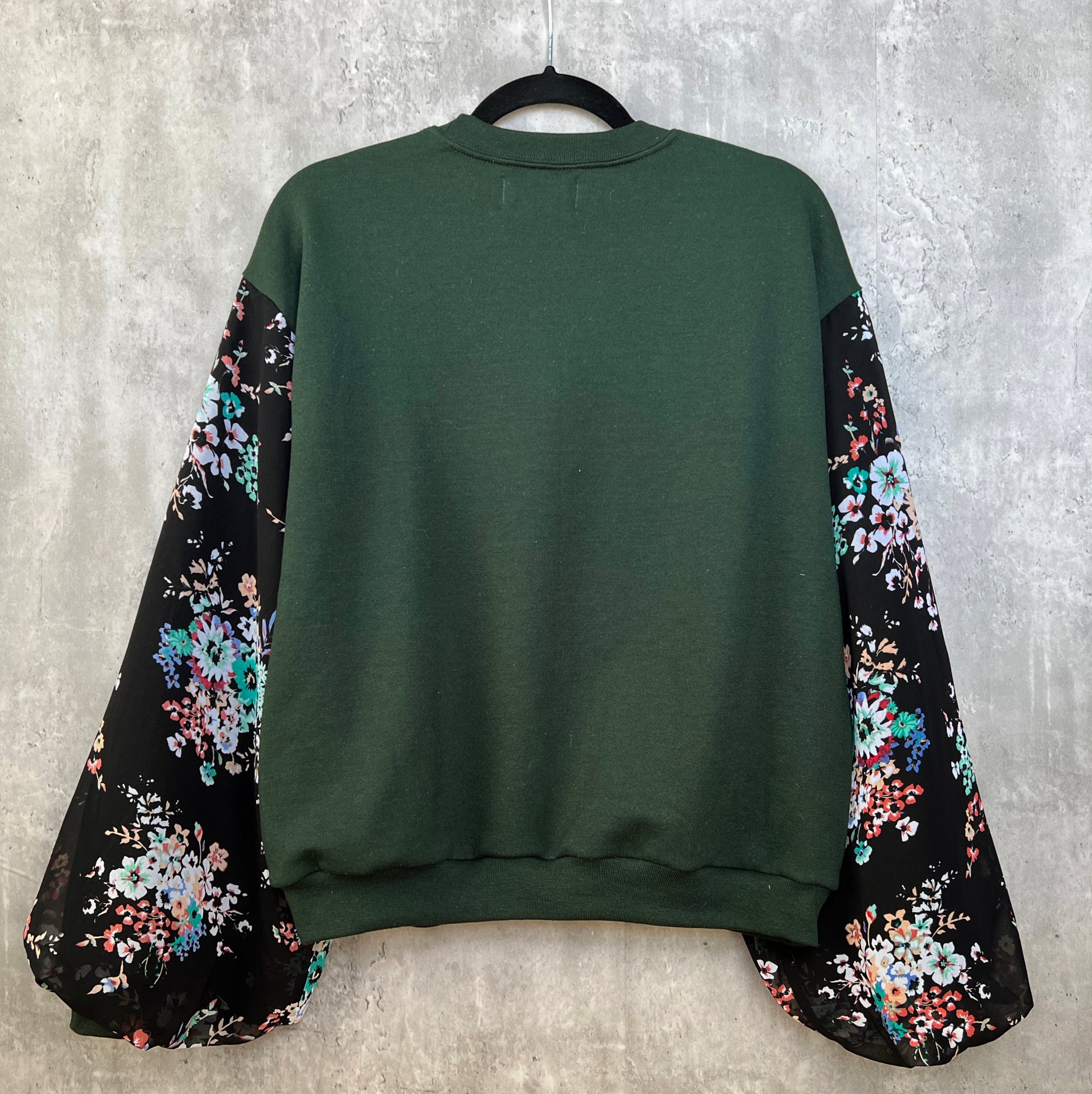 Back view of dark forest emerald green sweatshirt on hanger in front of cement gray background. Sweatshirt has puff sleeves with black base colorful floral. 
