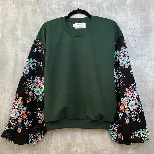 Front view of dark forest emerald green sweatshirt on hanger in front of cement gray background. Sweatshirt has puff sleeves with black base colorful floral. 